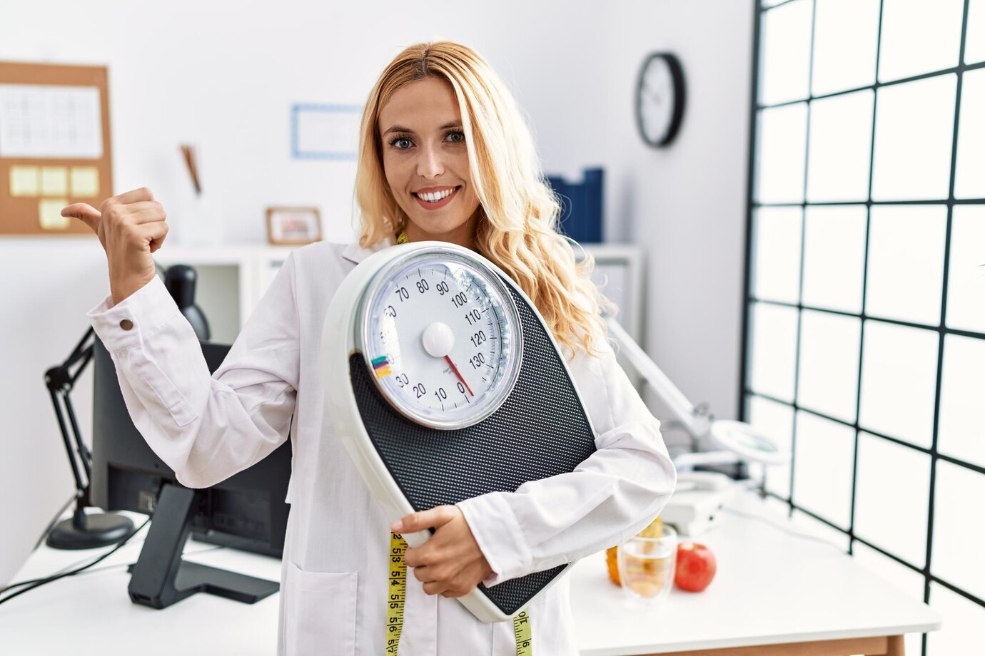 beautiful-blonde-nutritionist-woman-holding-weight-machine-balance-weight-loss-pointing-thumb-up-side-smiling-happy-with-open-mouth_839833-7437
